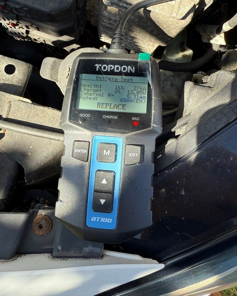 Battery tester in an engine bay reading 16 percent health and REPLACE, with the red BAD light on